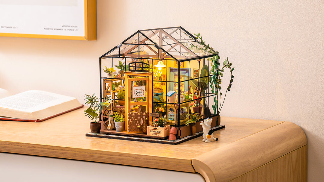 Robotime DIY craft product lifestyle image showing a completed miniature greenhouse model on a wooden desk with small plant and open book in a warm home setting