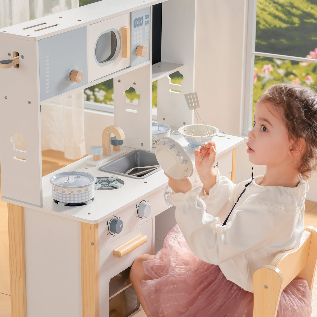 Girl seated beside the ROBUD white wooden play kitchen lifts a small cup while a pot rests on the stovetop burner.