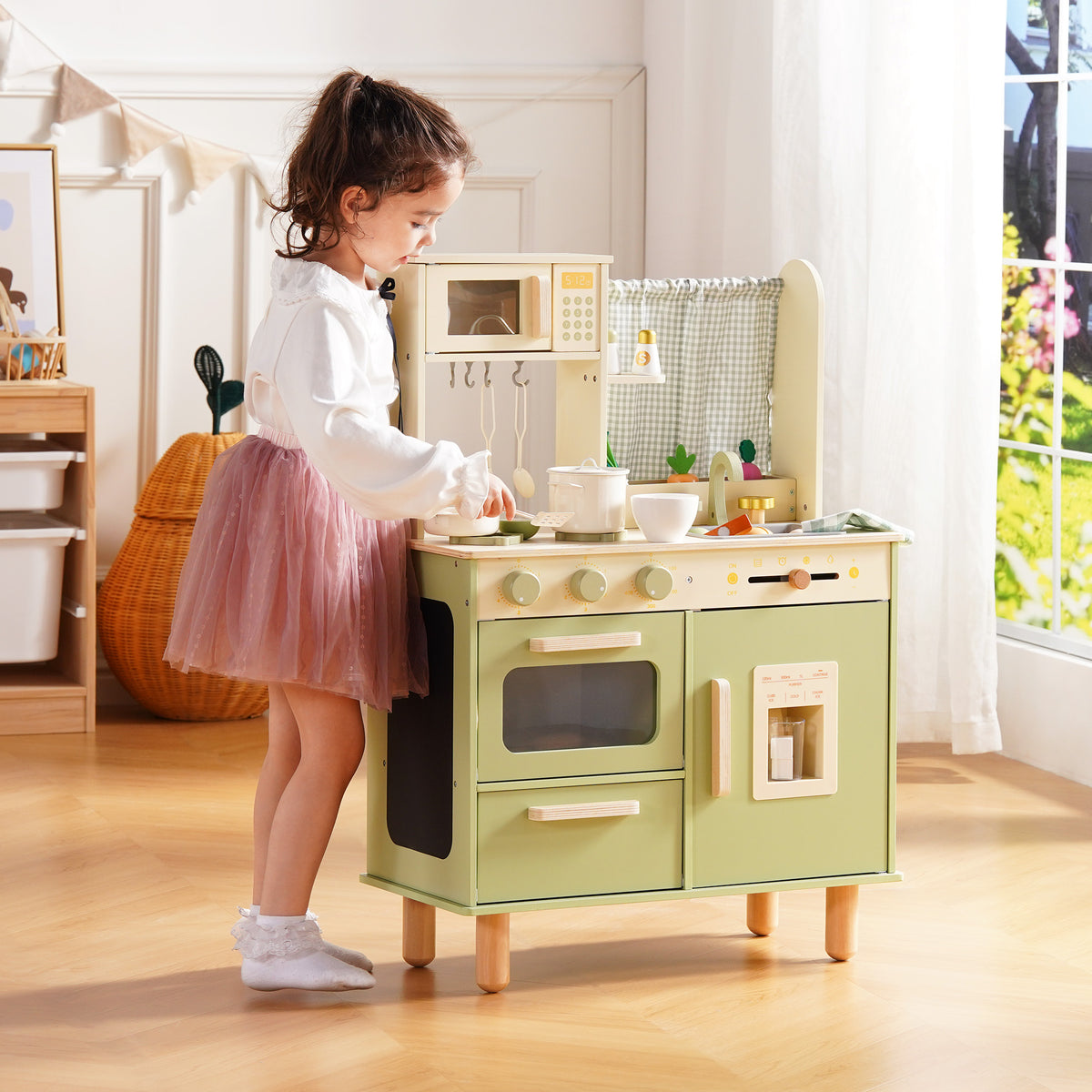 Girl in a pink tutu stands at the ROBUD sage green wooden play kitchen and opens the refrigerator door compartment.