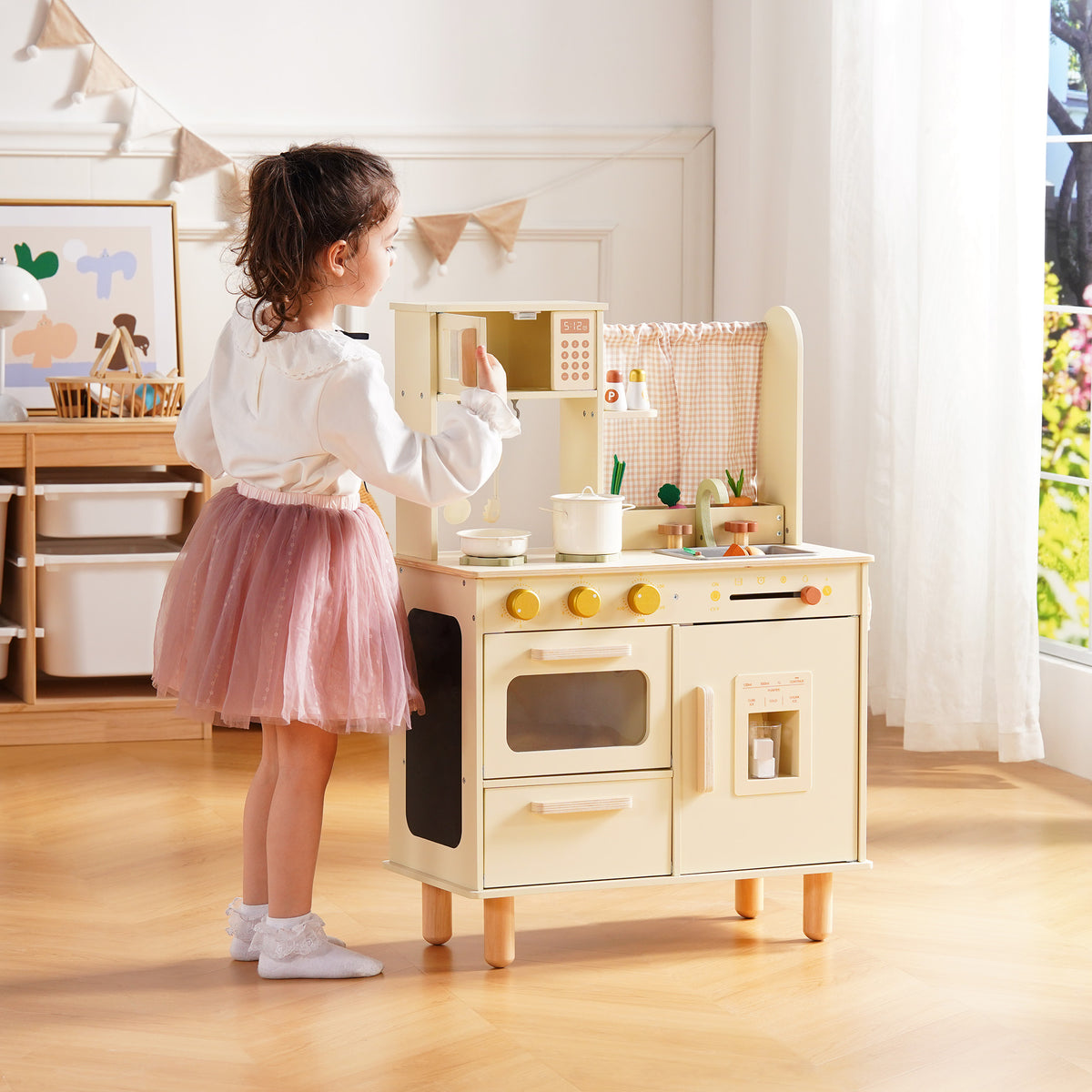 Girl in a pink tutu stands at the ROBUD cream wooden play kitchen, reaching toward the oven and refrigerator from the side.