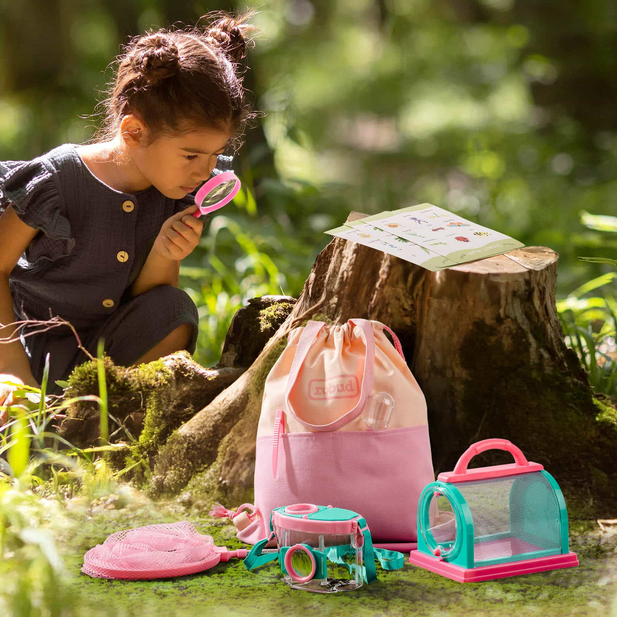 Young girl crouching in a woodland setting using a magnifying glass from the ROBUD pink outdoor explorer kit, with bug cage, magnifying jar, and leaf guide book on a log nearby
