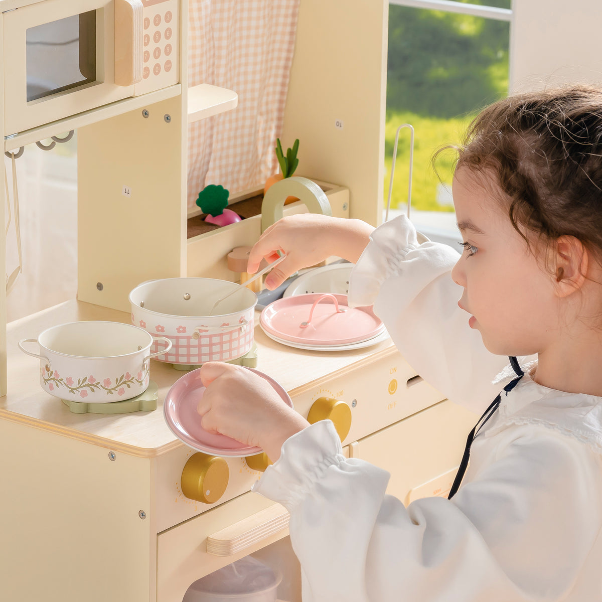 Girl in pink dress playing at wooden play kitchen with ROBUD Tinplate Pretend Cooking Utensils pink pots and food accessories on counter