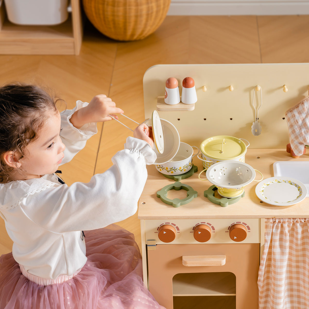 Girl pretend cooking with tongs at wooden kitchen toy while ROBUD Tinplate Pretend Cooking Utensils pink pots and plates are arranged on counter