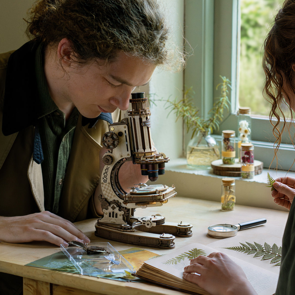 Two people examining botanical specimens on slides under the ROKR wooden microscope at a sunlit window ledge