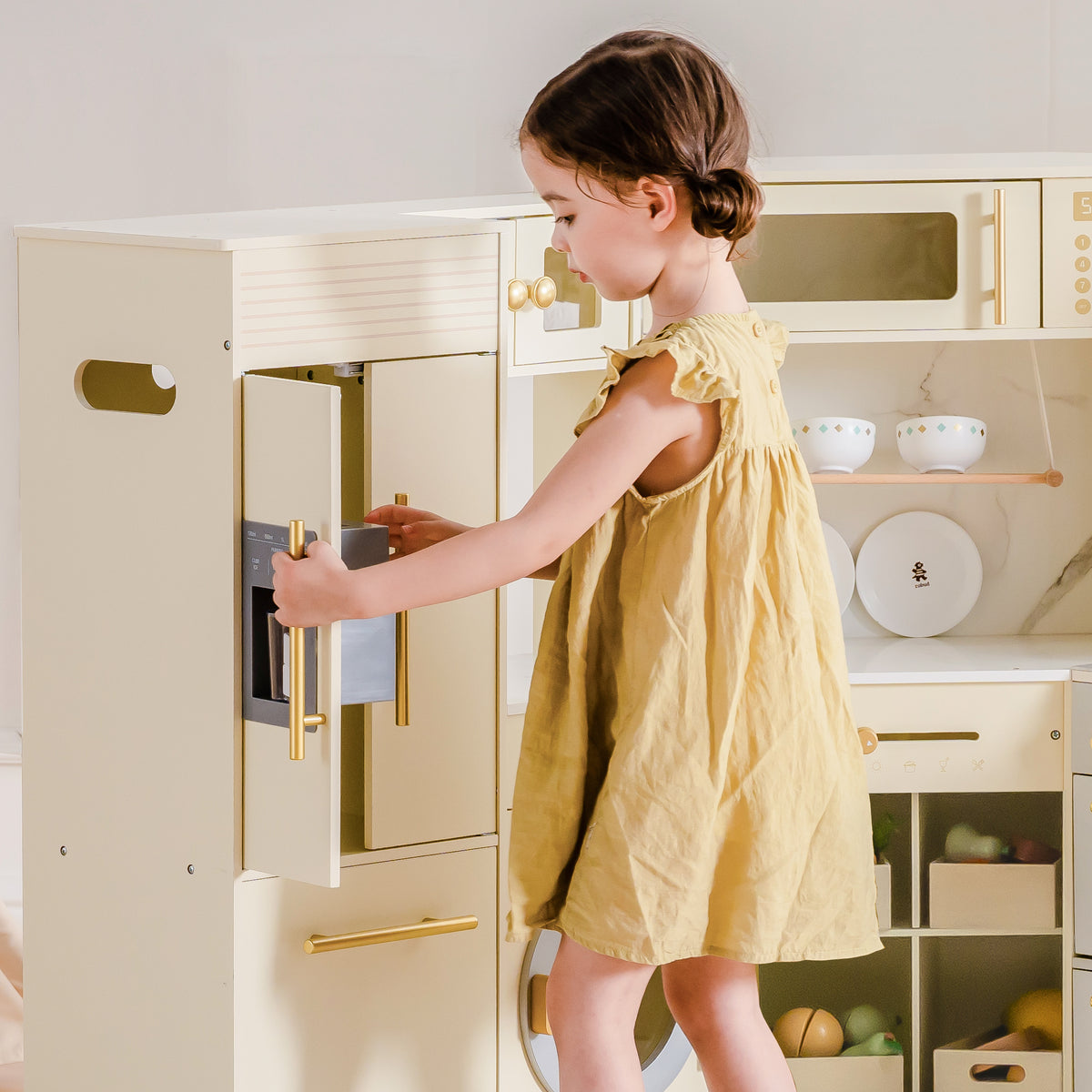 Young child in yellow dress opening the refrigerator door of ROBUD Corner Play Kitchen in cream and gold finish