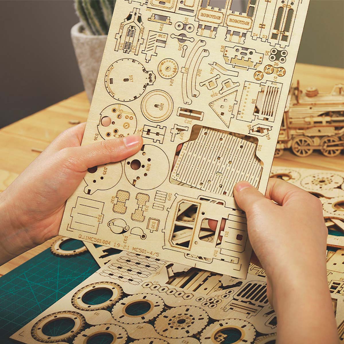 Hands holding a laser-cut wooden parts sheet from ROKR Army Jeep kit showing gears and body panel pieces