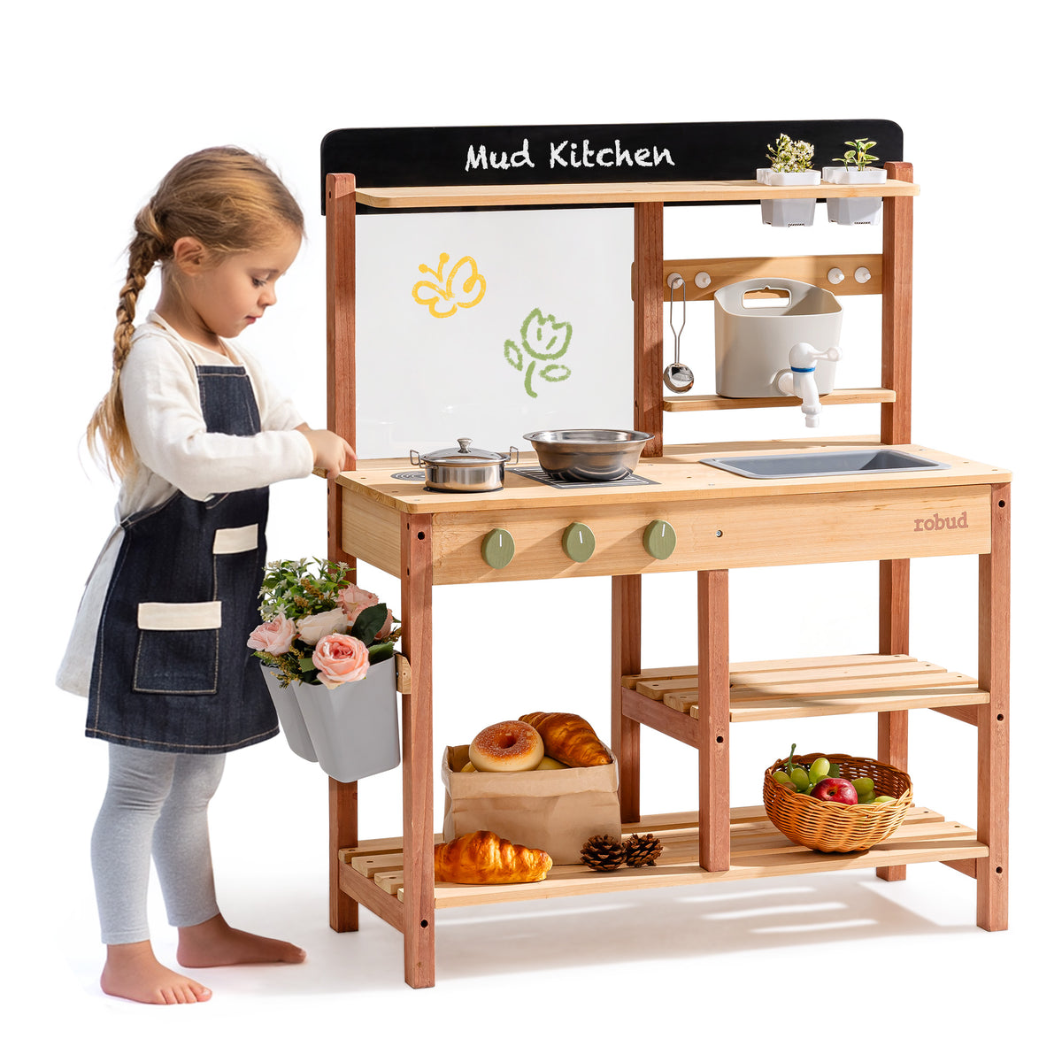 Girl in apron playing with the ROBUD outdoor wooden mud kitchen carrying a flower bucket, with baked goods and produce on the storage shelves