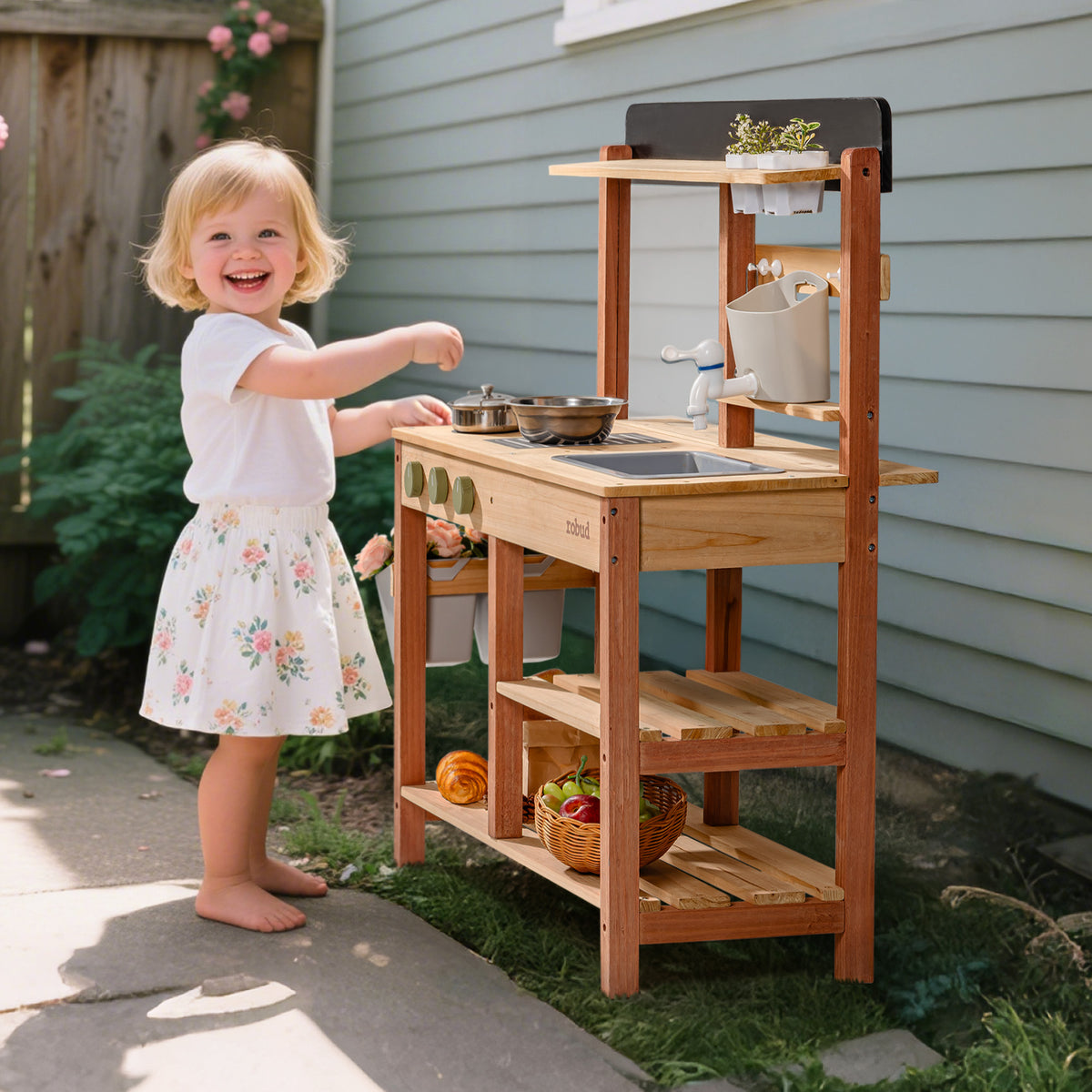 Smiling toddler girl outdoors beside the ROBUD wooden mud kitchen turning the tap while sunlight shines on the natural wood frame and storage shelves
