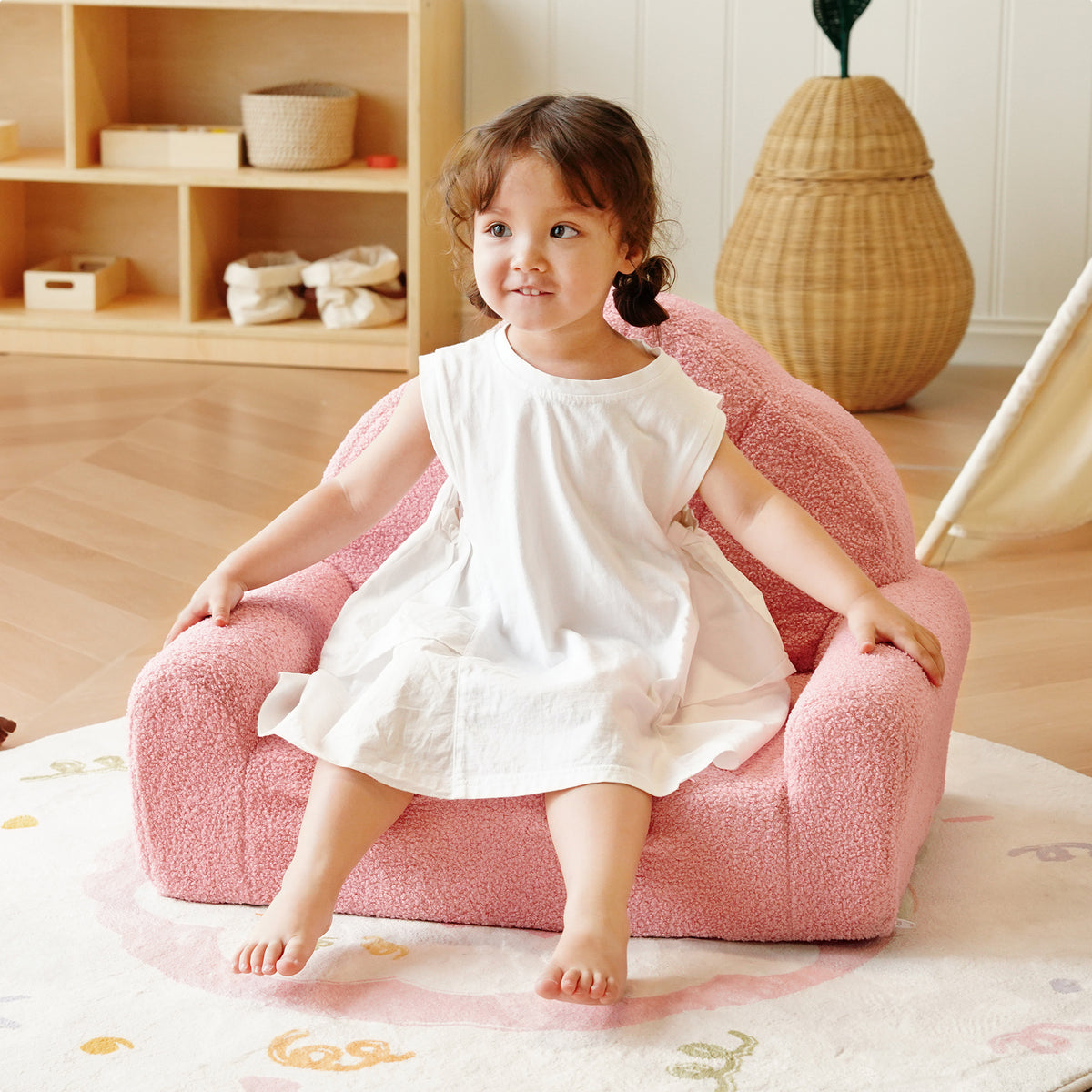 Young girl seated on the ROBUD pink Children's Sofa in a warm Scandi-style playroom with rattan basket