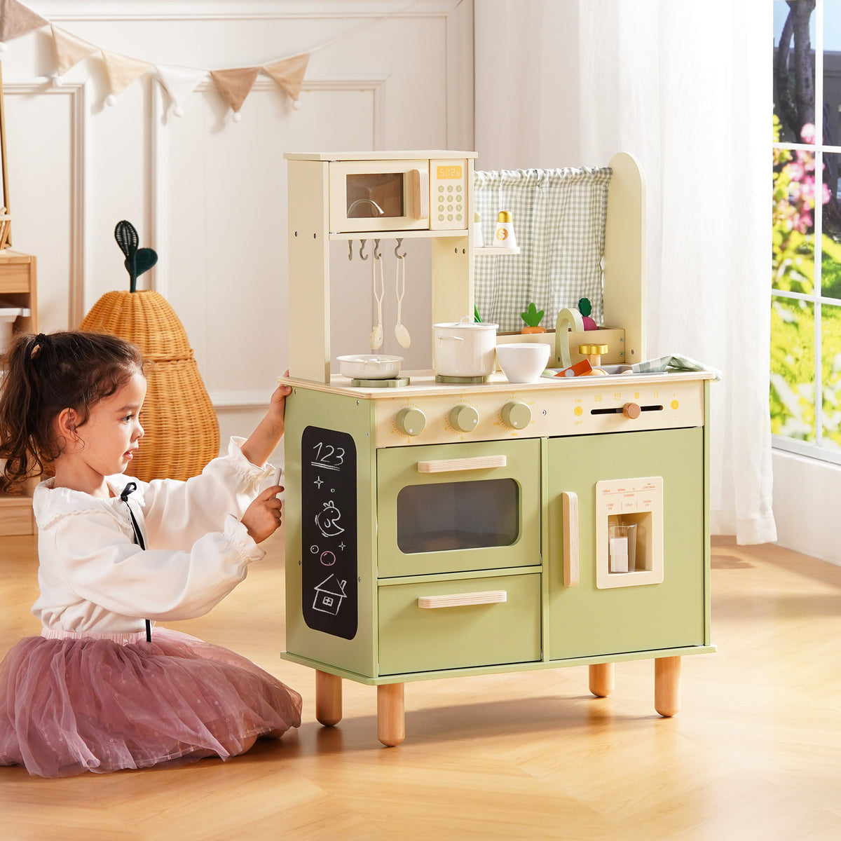 Girl in a tutu kneels beside the ROBUD sage green wooden play kitchen, writing on the chalkboard panel.