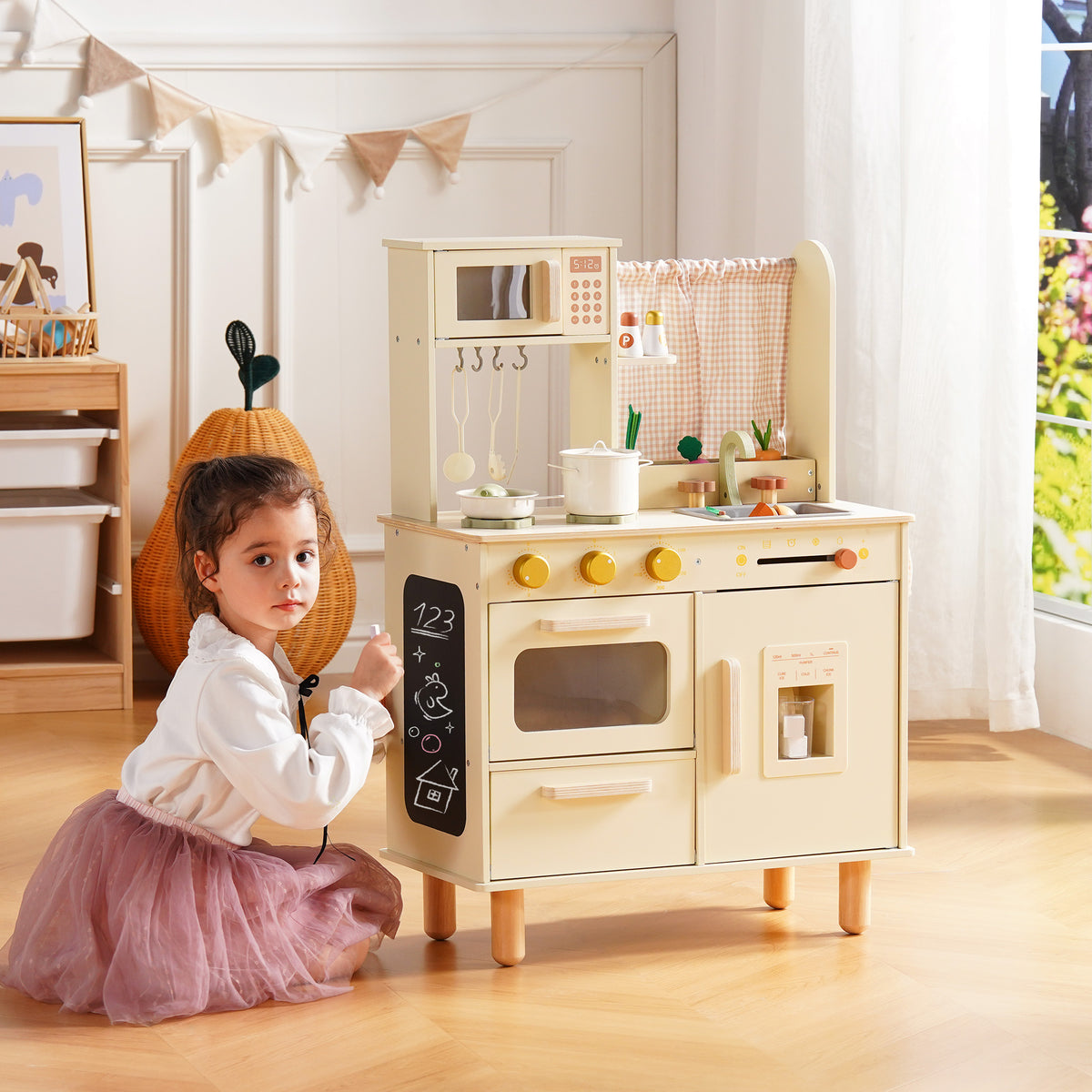 Girl in a tutu kneels at floor level beside the ROBUD cream wooden play kitchen, scribbling on the lower chalkboard panel.