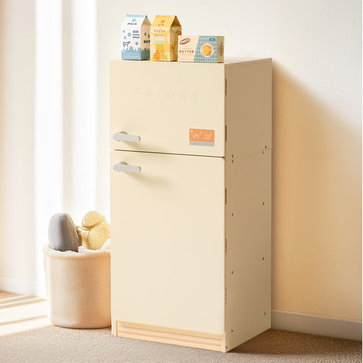 ROBUD creamy wooden play fridge styled in a warm room next to a basket, showing closed exterior with milk, juice, and butter cartons placed on top