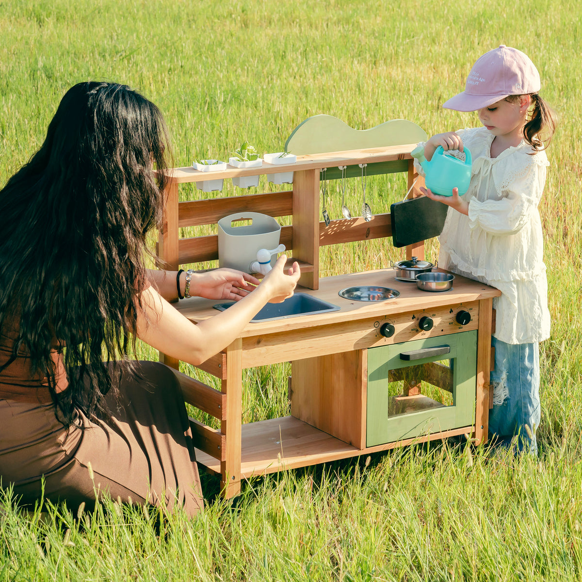 ROBUD Outdoor Mud Play Kitchen lifestyle view with mother sitting and girl playing beside the full honey-wood outdoor kitchen in lush green garden setting