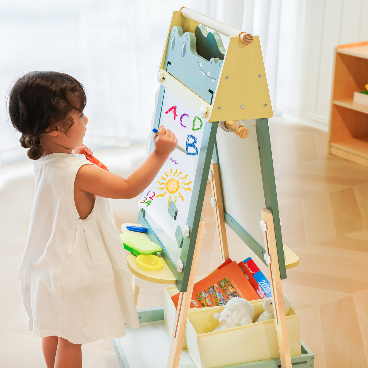 Toddler drawing letters on ROBUD Crocodile Easel in a sunny room with plants and toy guitar in background