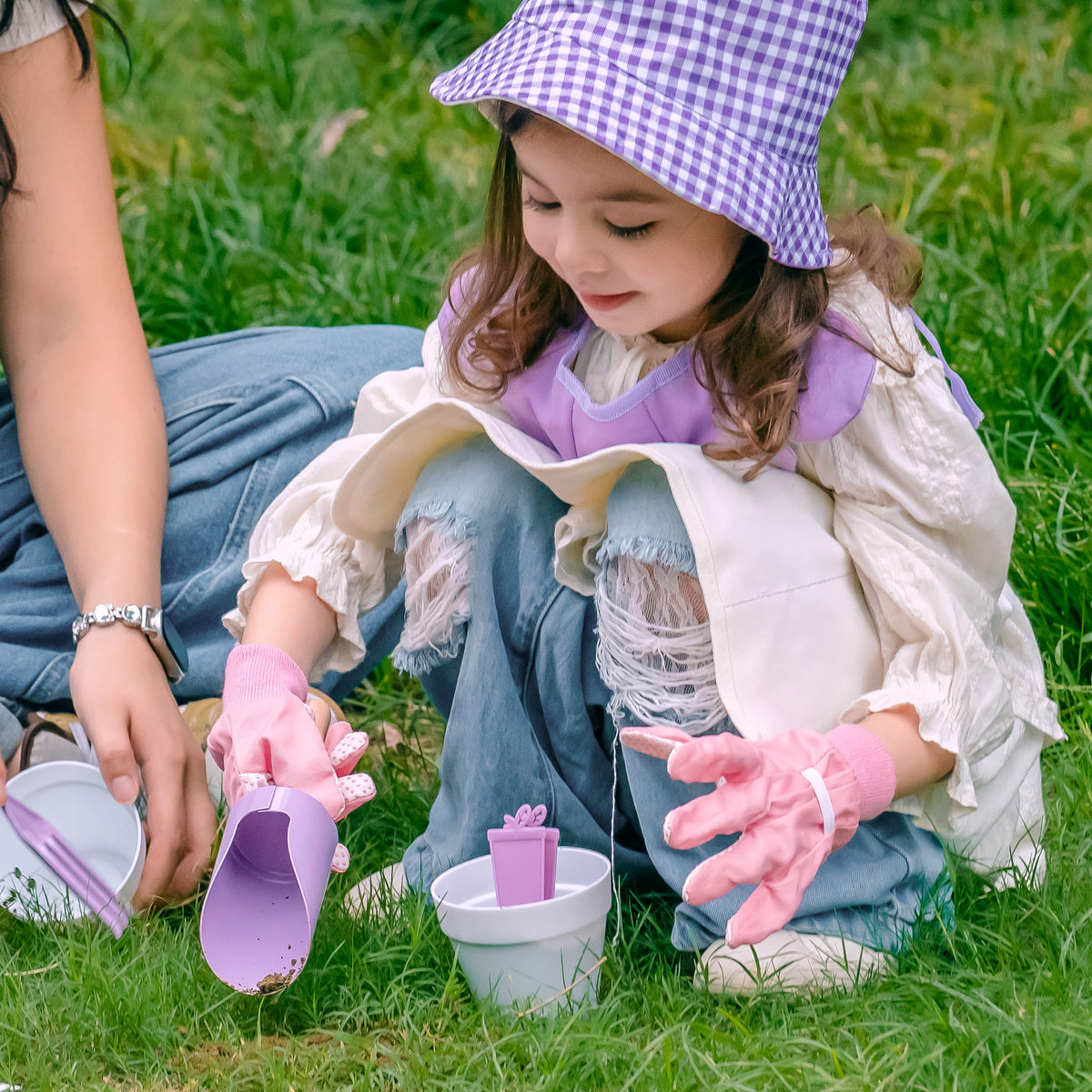 Girl in purple hat peering through ROBUD Kids Gardening Set magnifying glass outdoors among green plants and flowers
