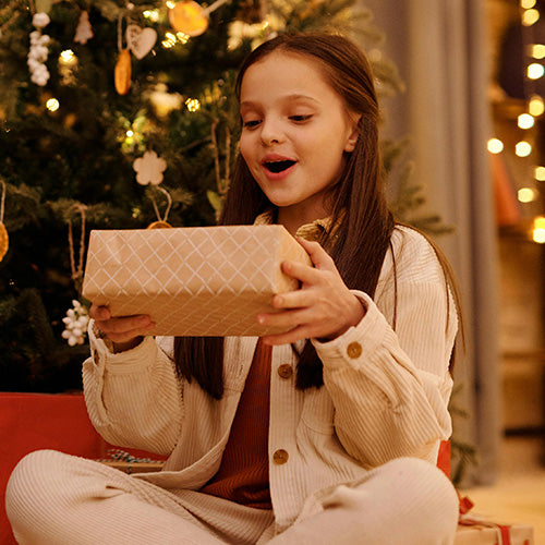 Excited young girl opening a wrapped gift box in front of a glowing Christmas tree, celebrating a holiday present moment