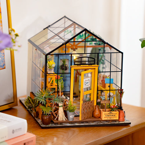 Rolife miniature greenhouse dollhouse with a yellow door, potted plants, garden tools and warm interior lighting displayed on a wooden desk