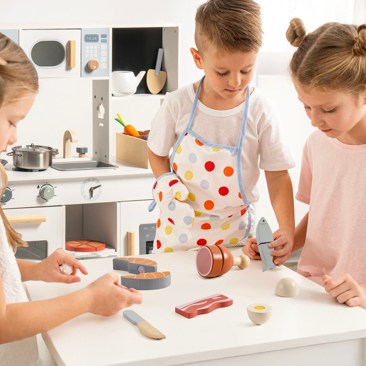 Three children at a white play kitchen table using ROBUD wooden meat cutting pieces including salmon, fish, bacon, and egg in pretend cooking play