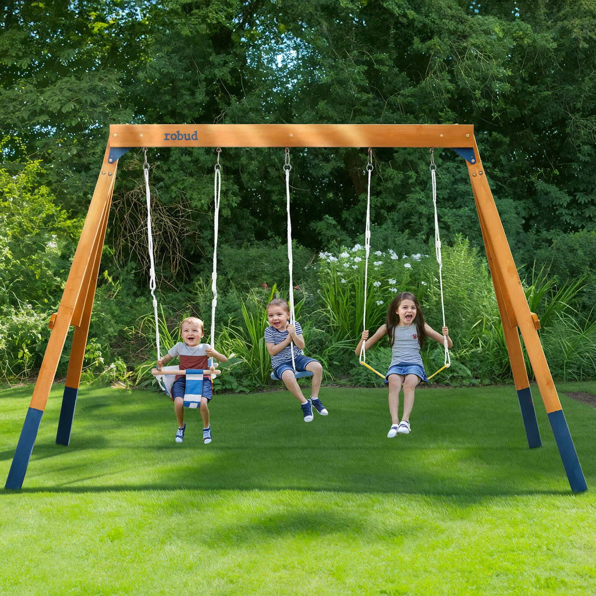 Three smiling children swinging on ROBUD natural wood A-frame outdoor swing set with yellow belt, blue disc and navy baby bucket seat among green trees