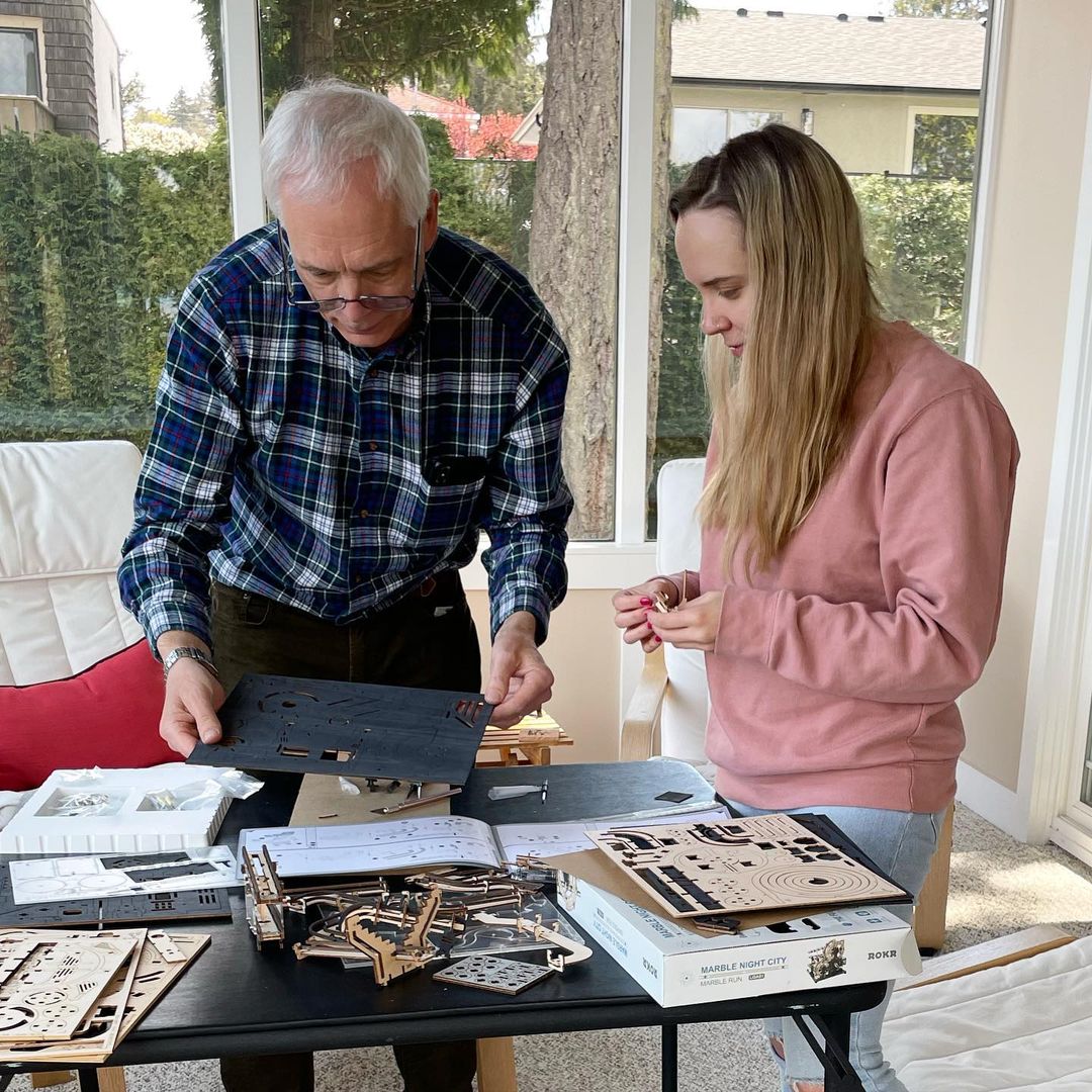 Man and young woman examining ROKR Marble Night City wooden puzzle sheets at a table covered with kit components