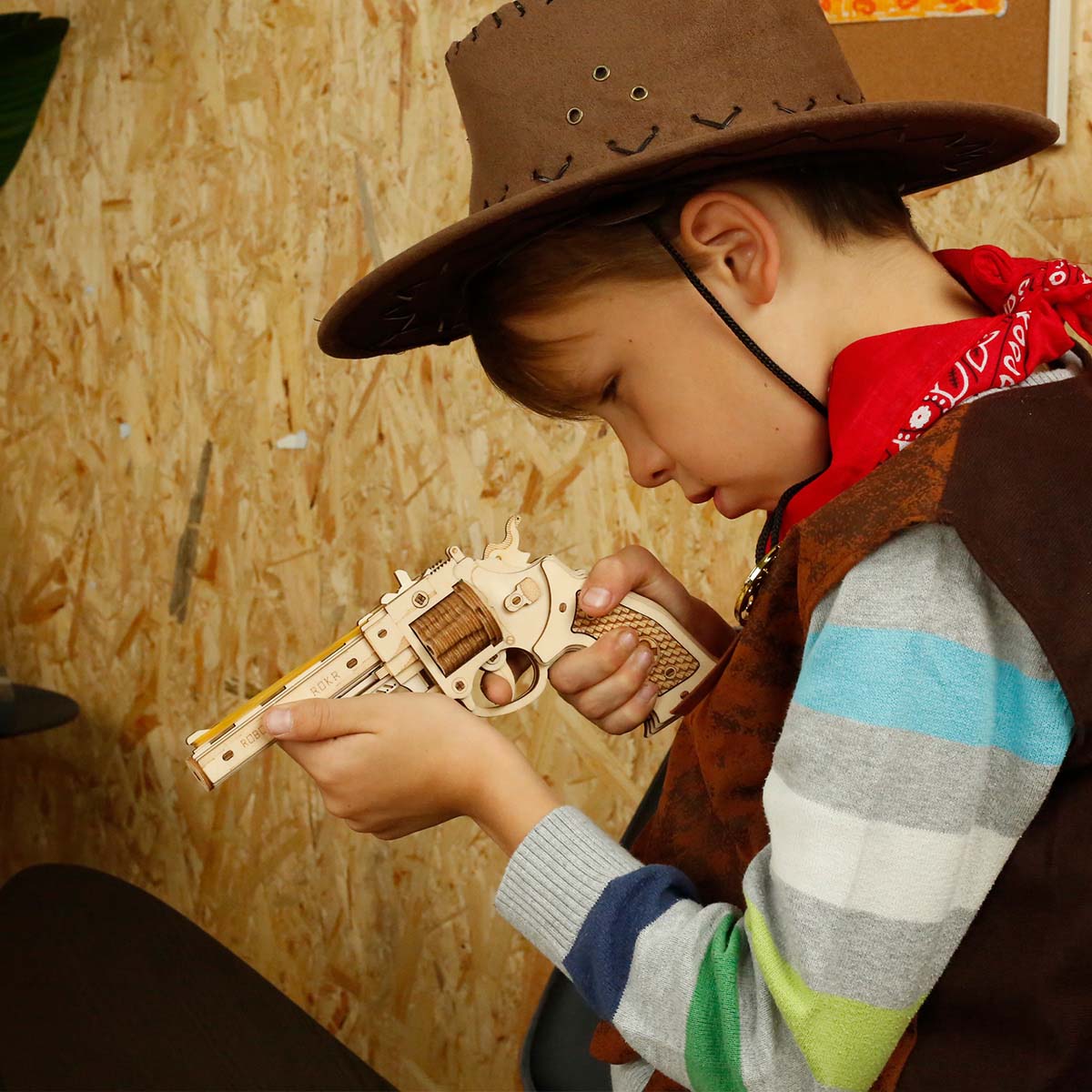 Boy in cowboy hat and brown vest closely inspecting a ROKR wooden revolver model against a plywood workshop wall