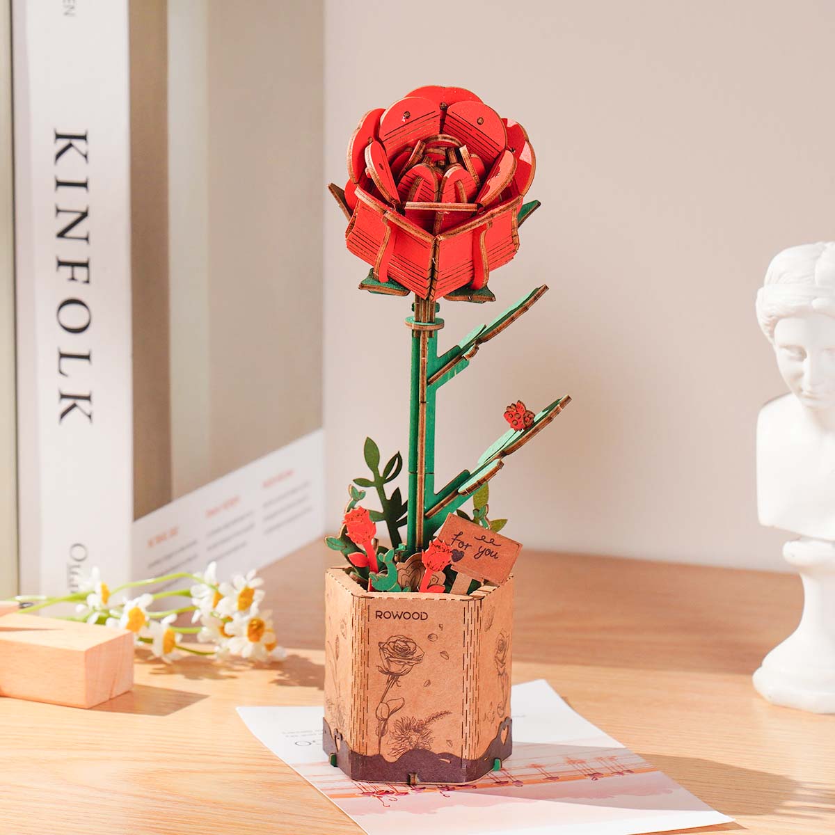 Rowood Red Rose 3D wooden puzzle standing in a kraft paper pot on a wooden desk beside a Kinfolk book and white plaster bust, with a tiny For You tag and green stem detail