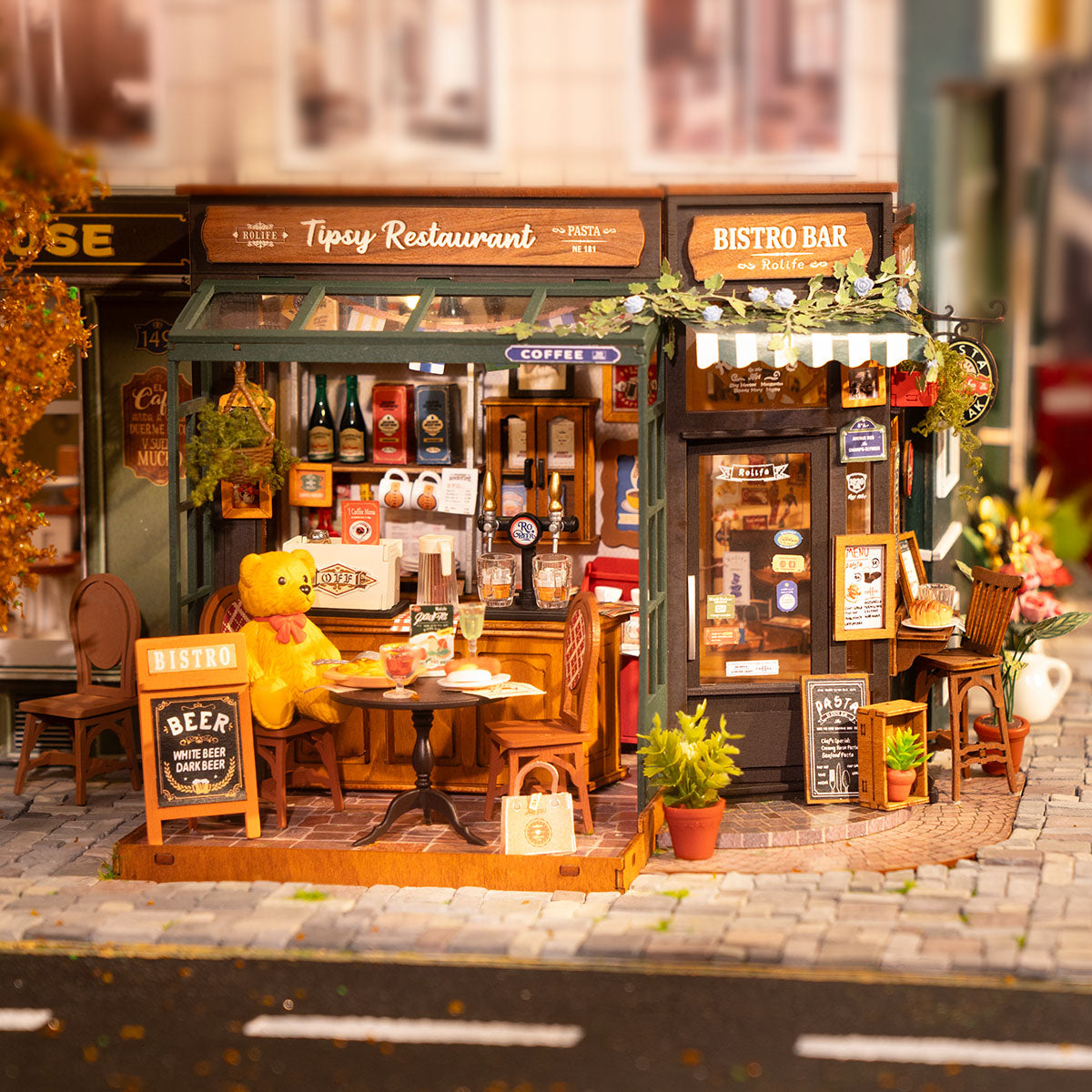 Rolife Tipsy Restaurant DIY miniature house DG167 displayed on a book stack with LED interior lights and a teddy bear seated at the cafe table
