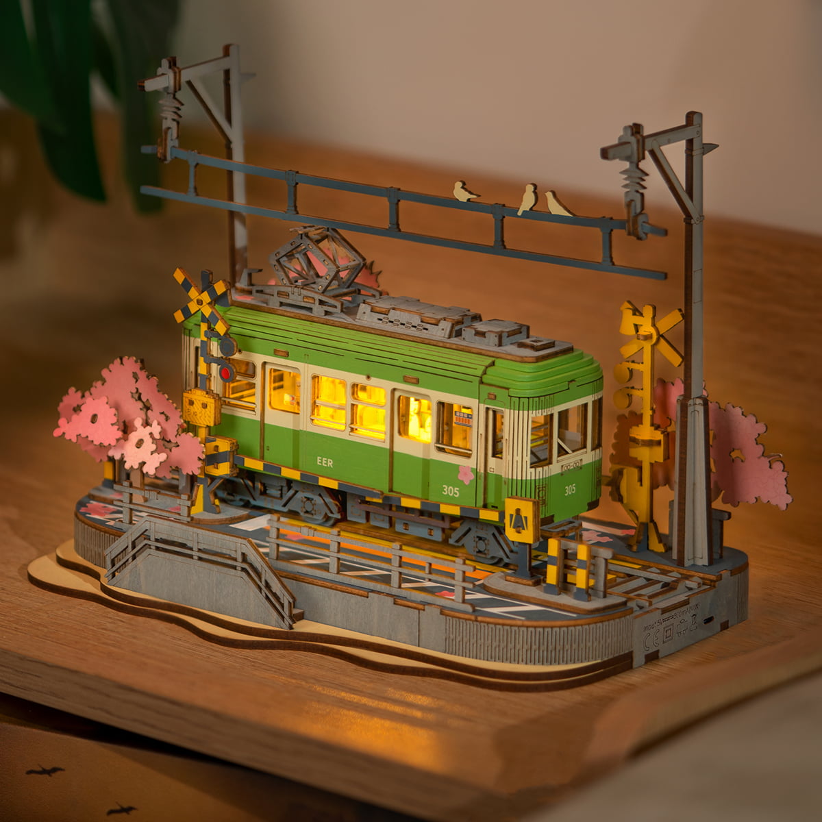 Rolife Sakura Journey tram model glowing with warm interior lighting amid yellow crossing signs and pink blossom trees
