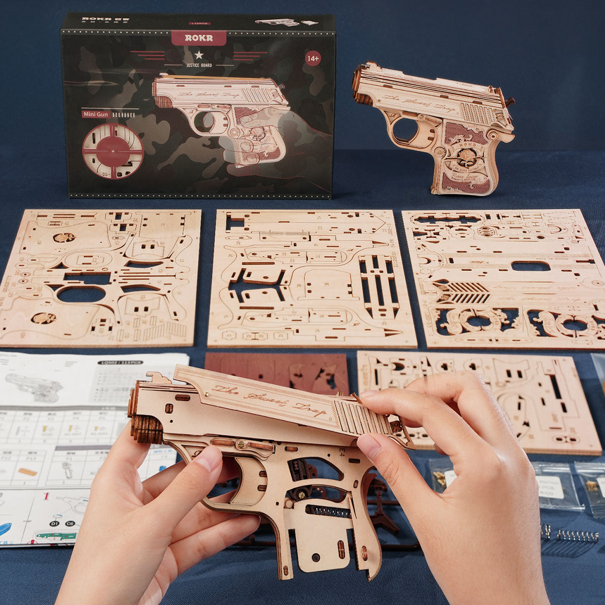 Hands assembling a ROKR wooden mini gun puzzle over instruction sheets and parts boards.