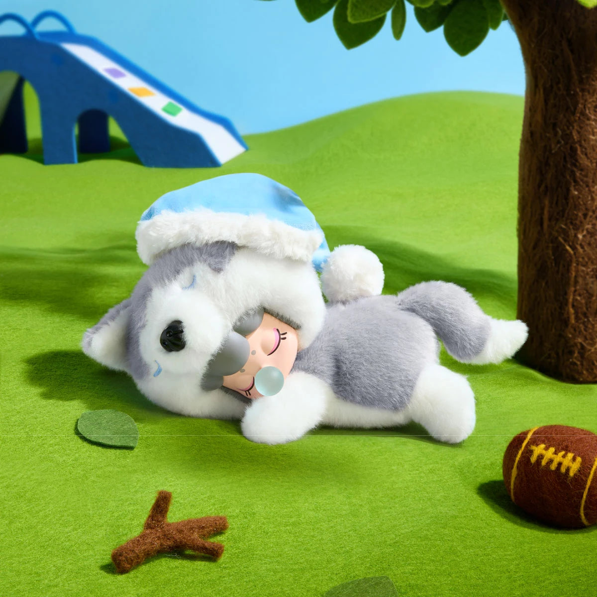 Grey and white plush husky toy with a doll face lying on green felt grass near a blue slide, tree, leaf, twig and brown football.