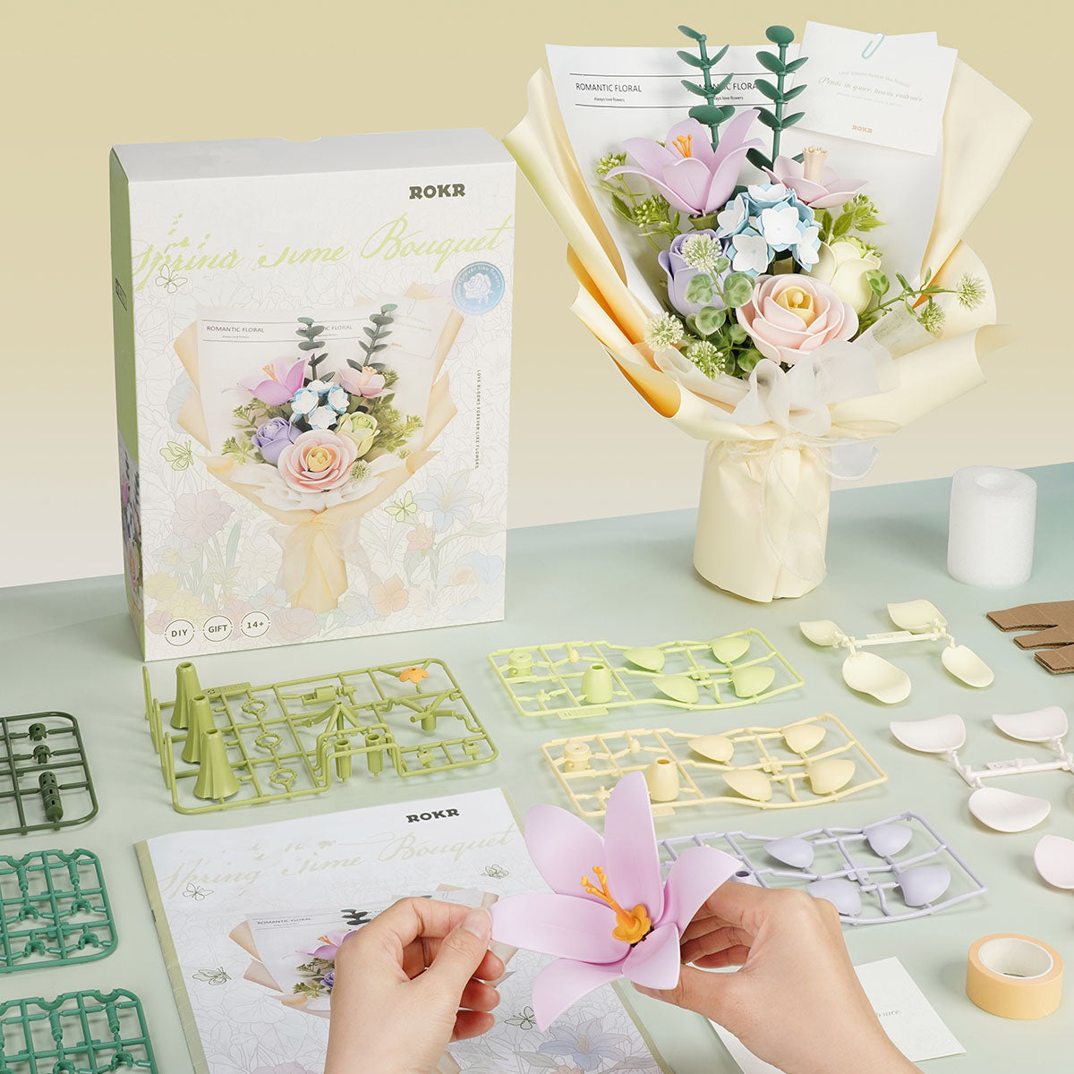 Hands assemble ROKR Spring Time Bouquet paper flower pieces on a desk with kit parts.