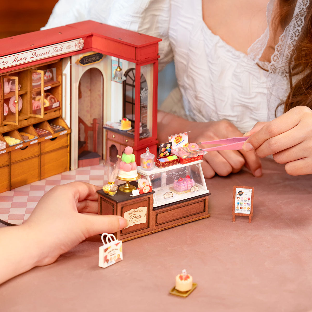 Hands placing miniature accessories beside Rolife Honey Dessert Talk DG168 dessert shop with cake and pastry details