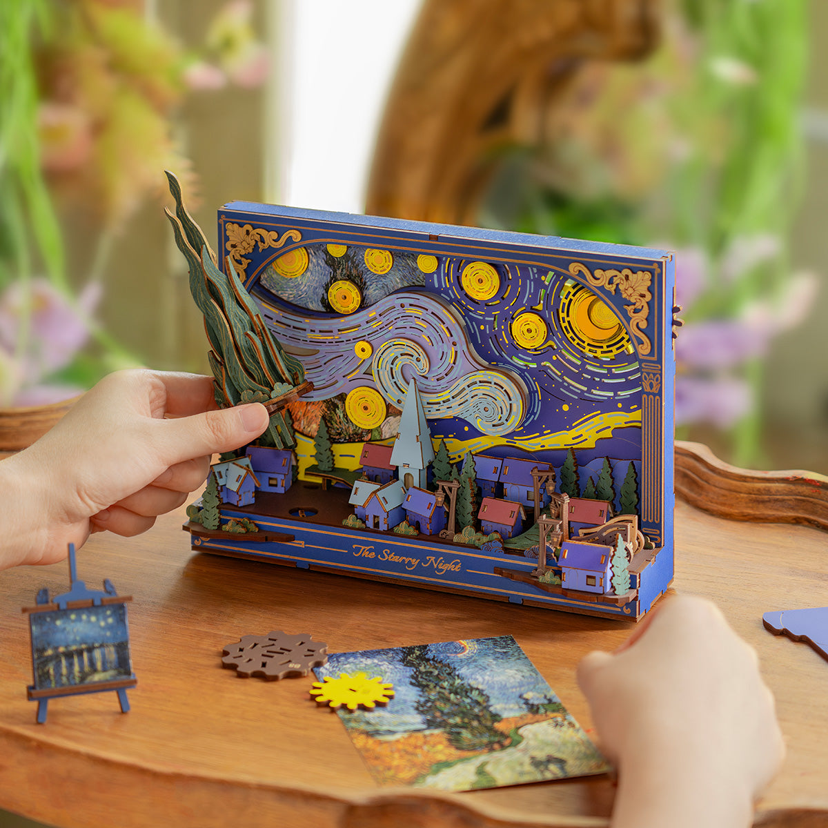 Rowood The Starry Night Wall Art 3D Wooden Puzzle TH003 lifestyle on a wooden tray near a painting, illuminated blue and gold Van Gogh scene glowing warmly beside an artist palette