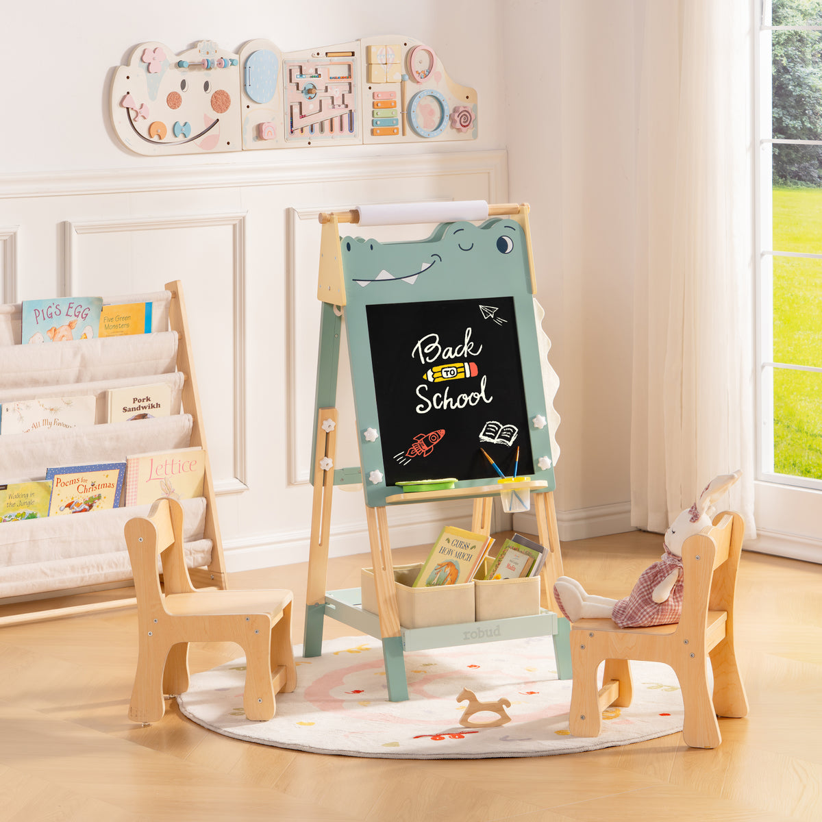 ROBUD green dinosaur easel with two small wooden chairs on a play mat surrounded by soft toys and books.