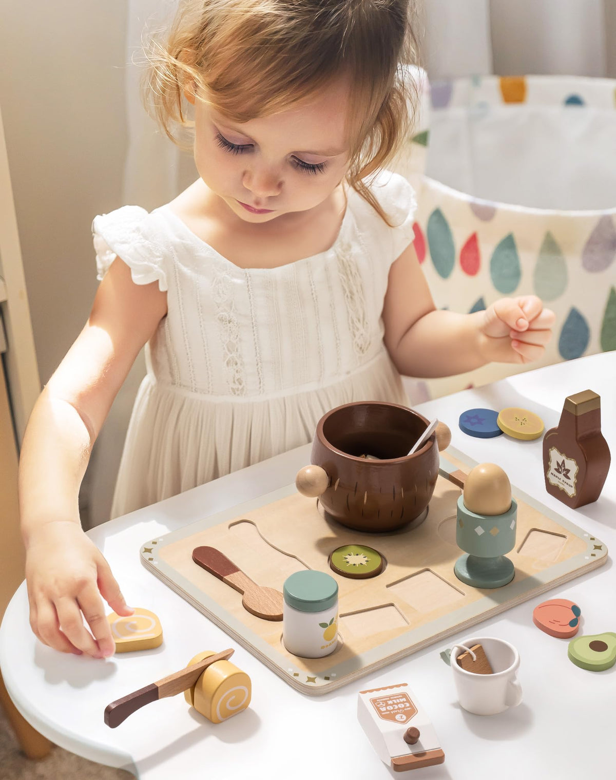 Young girl using ROBUD wooden food play set at white table with tray and natural wood kitchen nearby