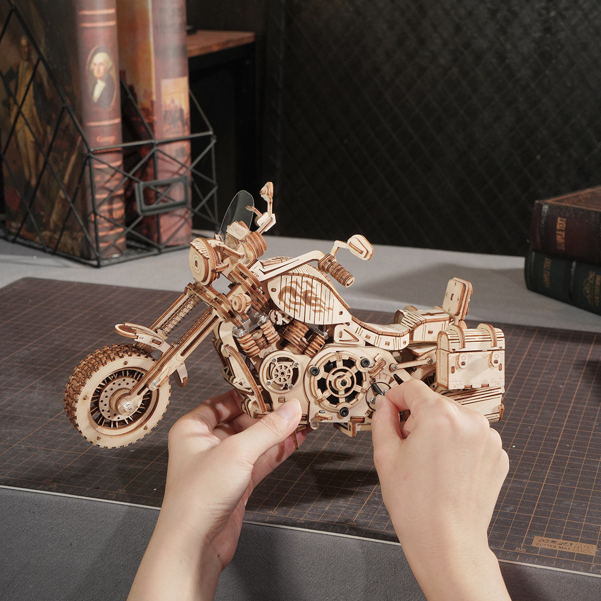 Hands placing a detail piece onto the assembled ROKR Cruiser Motorcycle wooden model beside stacked books