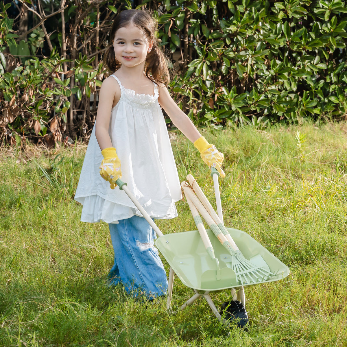 Smiling girl pushing sage green ROBUD Kids Wheelbarrow through a grassy garden wearing white dress and yellow gloves