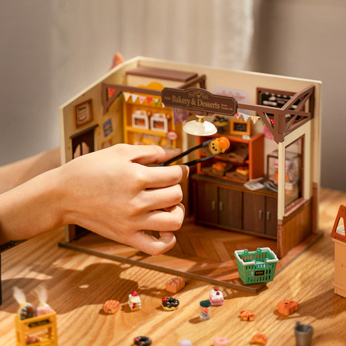 Hands assembling Rolife Beckas Baking House miniature bakery kit with scattered wooden parts and accessories on table