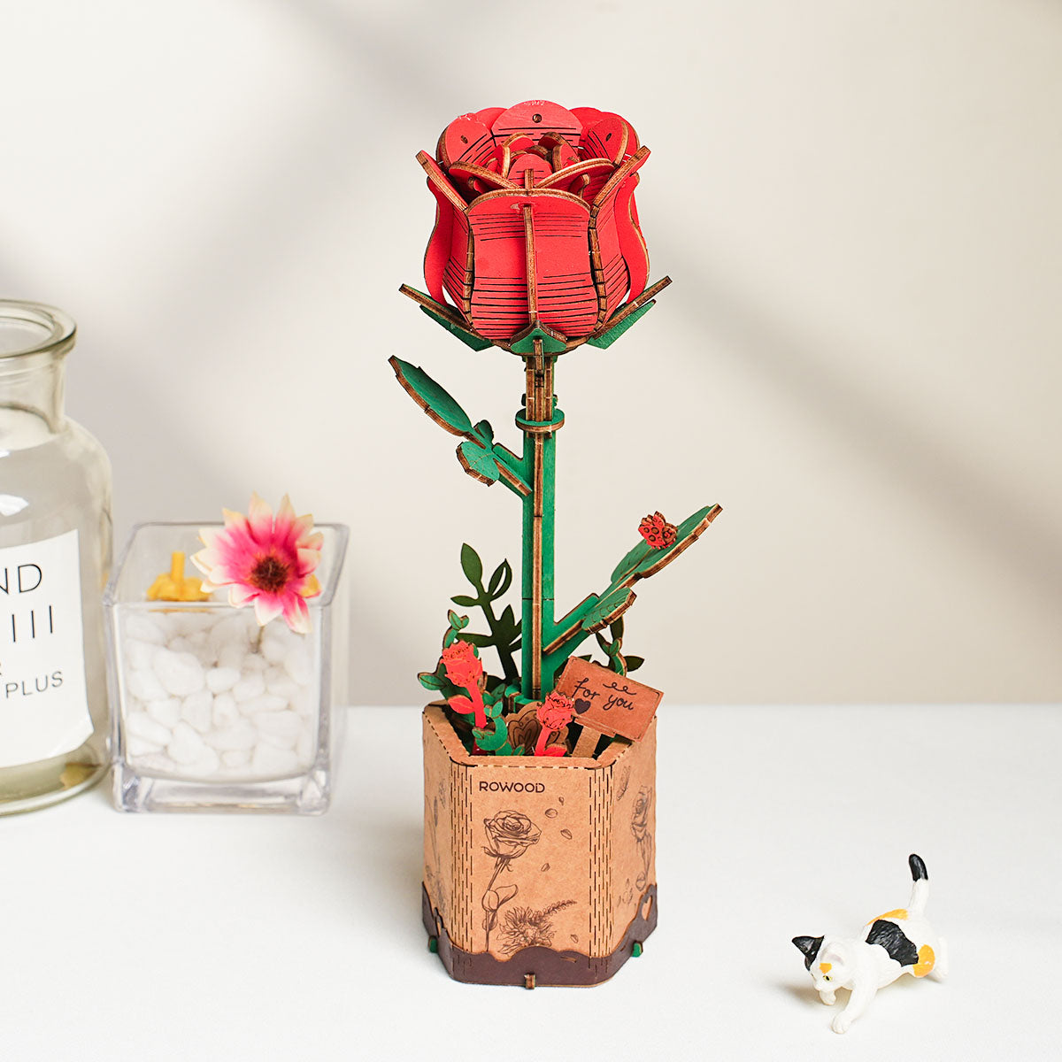 Rowood Red Rose 3D wooden puzzle in a kraft paper pot displayed beside a glass jar with pebbles and a small pink flower, with a black and white cat figurine nearby