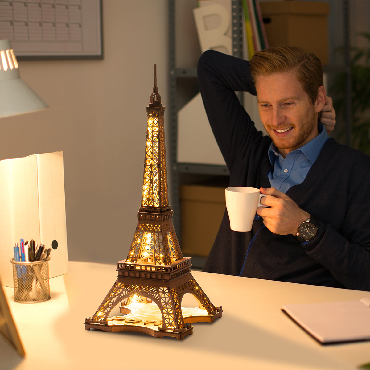 Man relaxing at a desk with a cup of coffee while a fully assembled Rolife Eiffel Tower wooden model glows with warm LED light