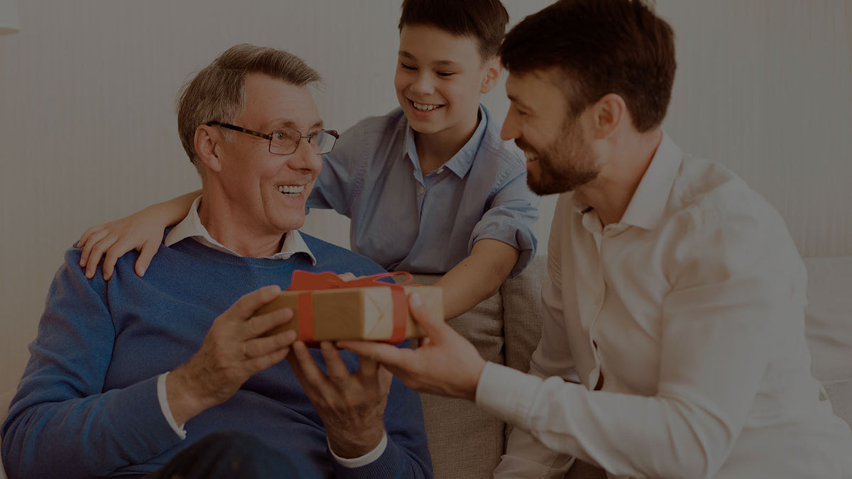 Dimly lit lifestyle banner showing a grandfather delightedly receiving a Robotime gift from a bearded son while a boy watches in a warm living room