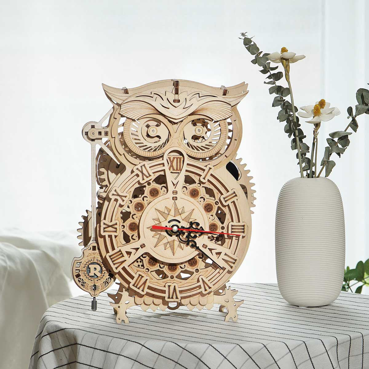ROKR Owl Clock LK503 on a striped linen tablecloth beside a white vase with eucalyptus, warm indoor setting