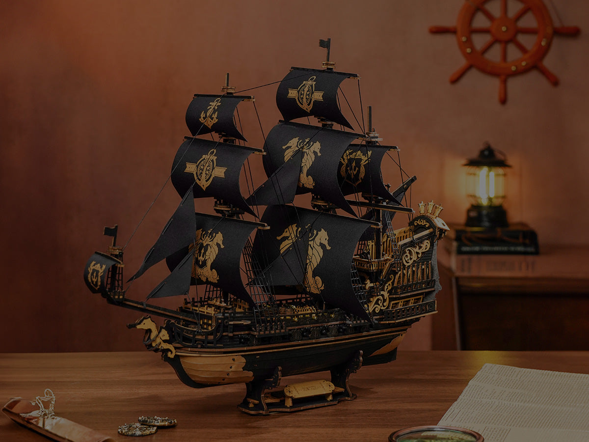 ROKR wooden pirate ship 3D puzzle model with black sails printed with golden seahorse and anchor motifs displayed on a desk with a ship wheel, compass and lantern in the background