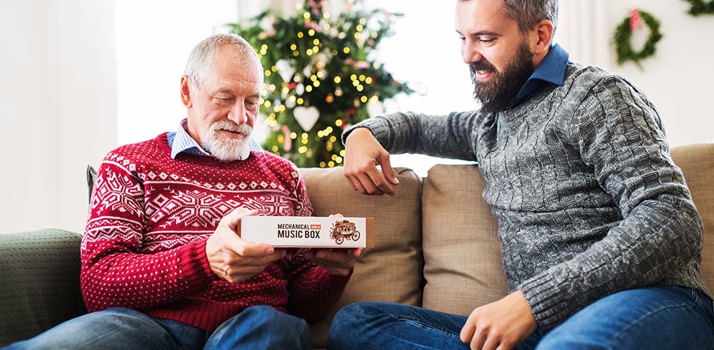 Two men smiling on a sofa near a Christmas tree, one holding a ROKR Mechanical Music Box package, holiday wreath visible in the background