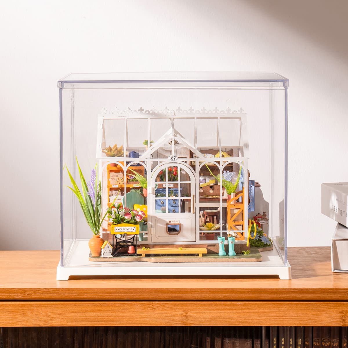 Rolife Dreamy Garden House miniature sits under a clear case on a wooden cabinet.
