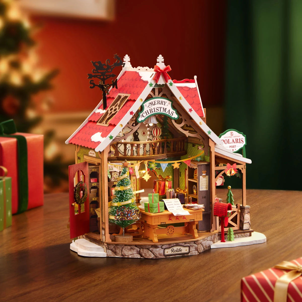 Rolife miniature Christmas post office model on a wooden table, showing open front, Santa at desk, decorated tree, red mailbox and Polaris Post sign, with wrapped gifts nearby.