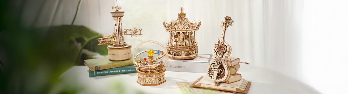 ROKR mechanical music box collection displayed on a desk featuring four models including a carousel, cello, orrery with crystal planets, and airplane tower in warm natural wood tones