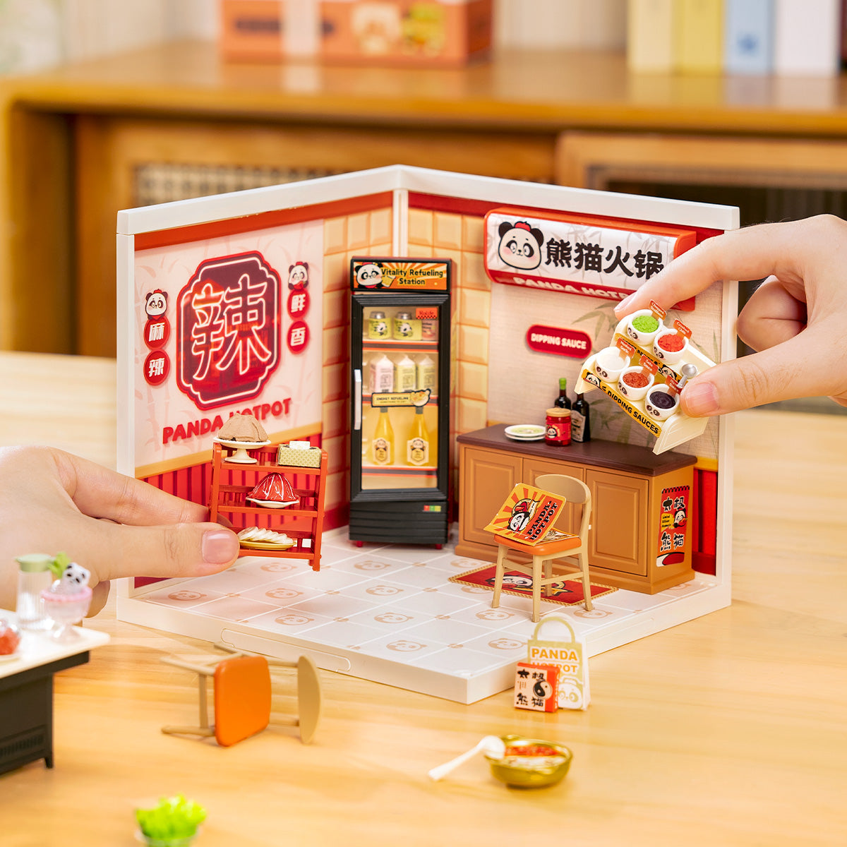 Hands opening the Rolife Panda Hot Pot DW020 Super Creator kit while a panda hotpot sign and illuminated drink fridge are visible inside the assembled room