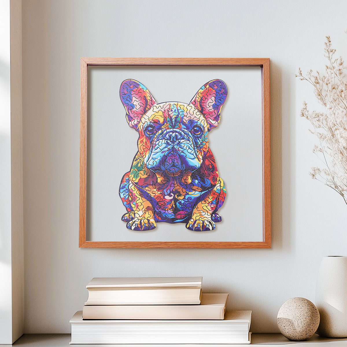 Rowood French Bulldog colorful wooden jigsaw puzzle in a shadow box frame hung on a wall above a stack of books and a sphere ornament