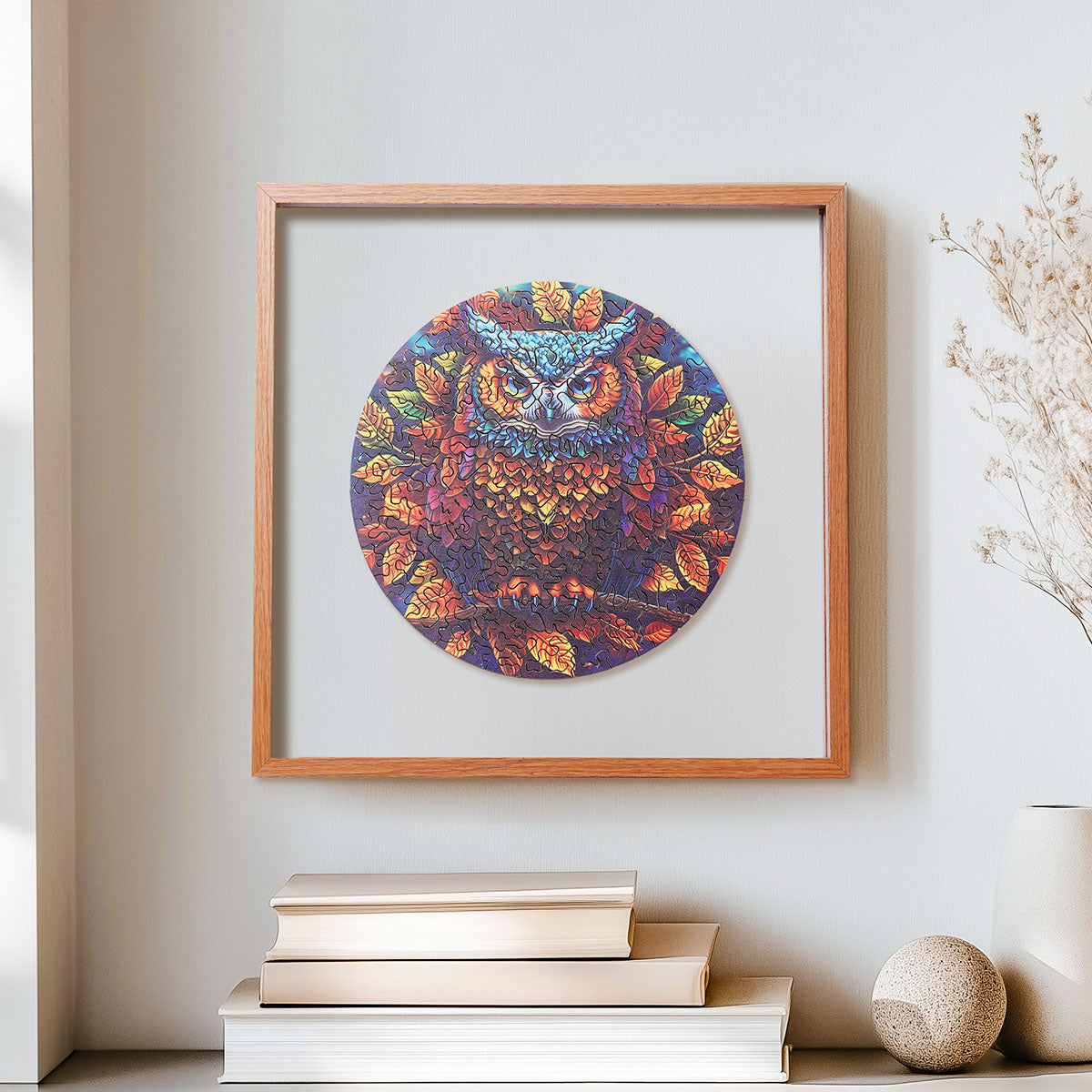 Rowood Moonlight Owl circular wooden jigsaw puzzle in a shadow box frame hanging above books on a wall, surrounded by dried pampas grass